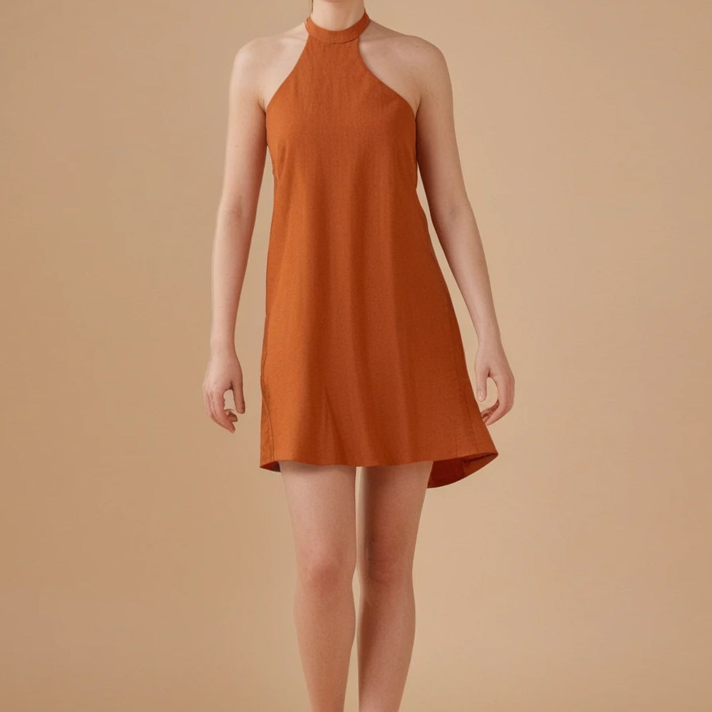 A woman wears a sleeveless, burnt orange halter-neck mini dress with an A-line silhouette. The dress features a high neckline and a relaxed fit that gently flares at the hem. The fabric appears lightweight, perfect for warm weather. The model stands against a beige background, highlighting the earthy tone of the dress. This minimalist design is part of a sewing pattern collection for summer dresses.