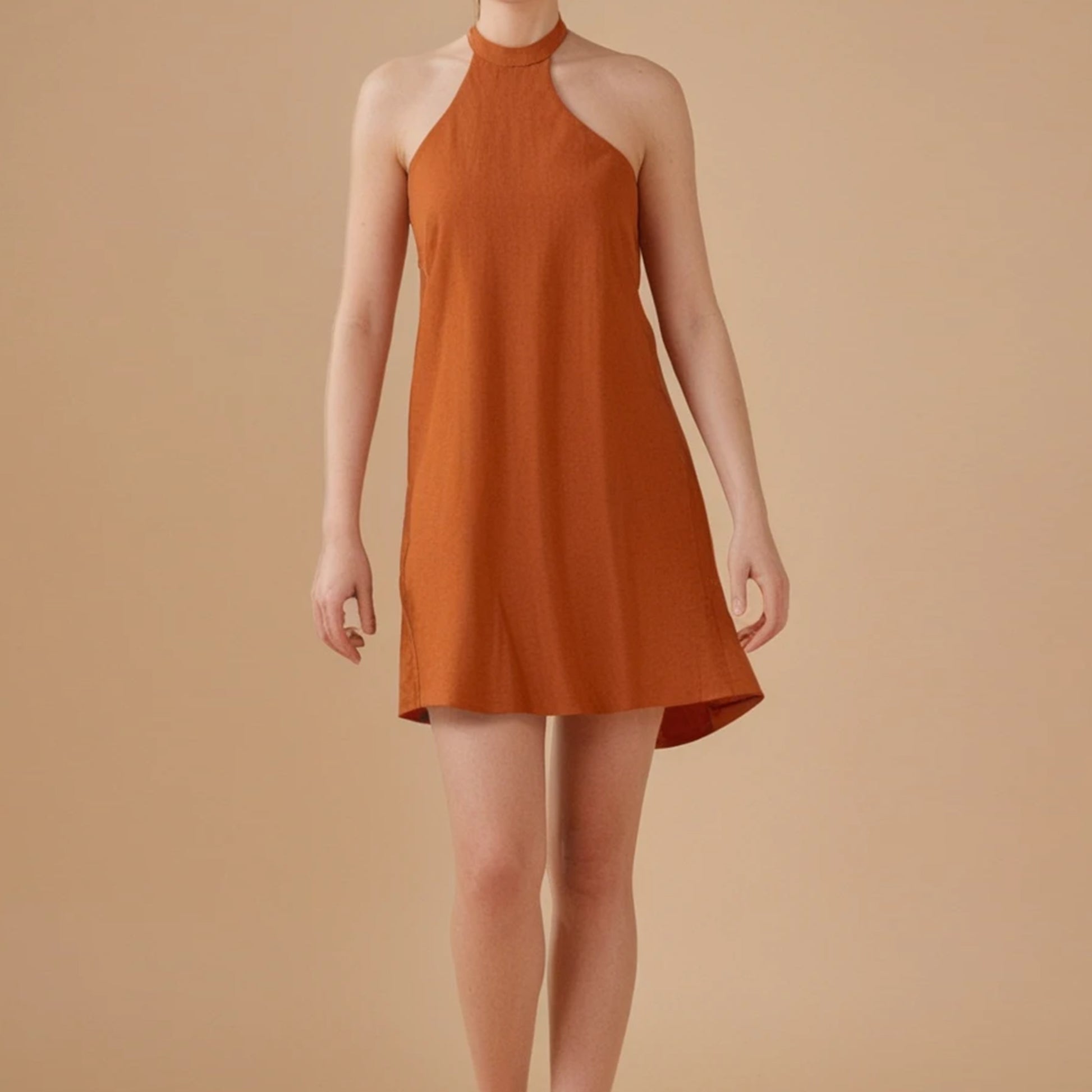 A woman wears a sleeveless, burnt orange halter-neck mini dress with an A-line silhouette. The dress features a high neckline and a relaxed fit that gently flares at the hem. The fabric appears lightweight, perfect for warm weather. The model stands against a beige background, highlighting the earthy tone of the dress. This minimalist design is part of a sewing pattern collection for summer dresses.