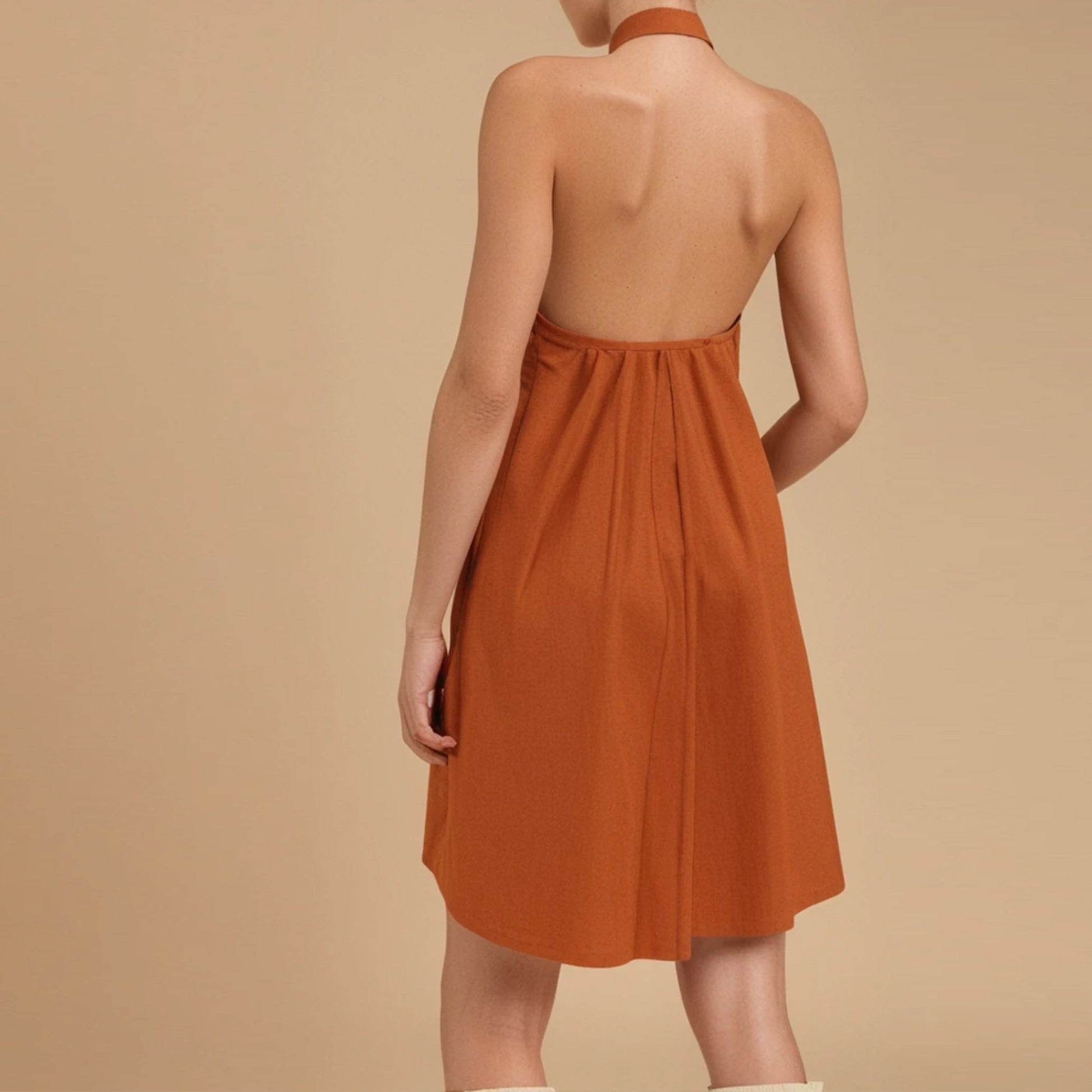 Rear view of a woman wearing a sleeveless, burnt orange halter-neck mini dress with a completely backless design. The dress gathers slightly at the back waist, adding a soft drape and ease of movement. The halter strap ties or fastens around the neck, leaving the shoulders and upper back fully exposed. The garment falls to above the knee and has a relaxed, A-line shape. The model stands against a plain beige background, showcasing a breezy, summer-appropriate sewing pattern.