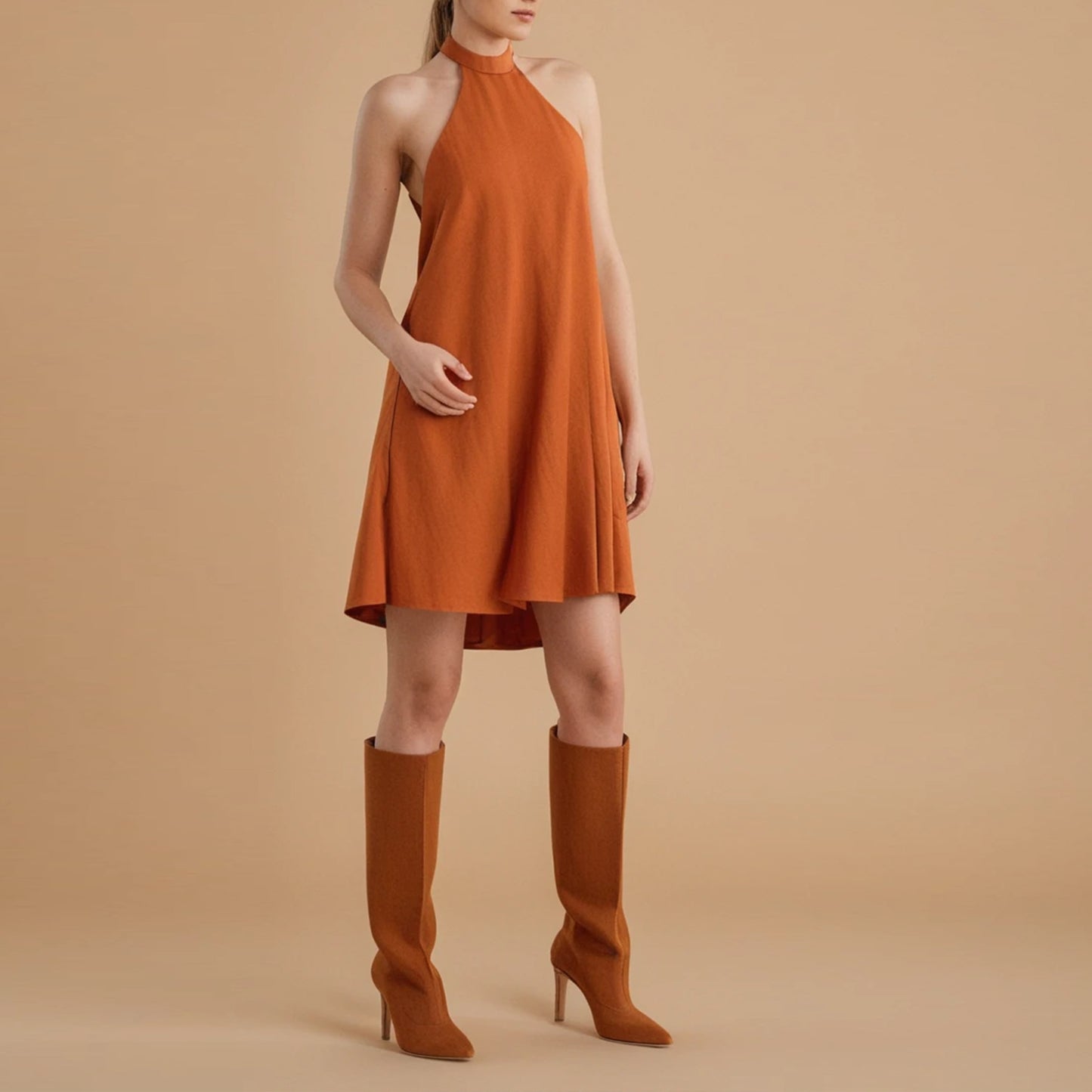 Front view of a woman wearing a sleeveless, burnt orange halter-neck mini dress with an A-line silhouette. The dress features a high neckline and a flowy, relaxed fit that flares out slightly toward the hem, creating an effortless and breezy look. The model pairs the dress with matching high-heeled, knee-high boots in a similar rust tone. She poses against a solid beige background, showcasing the minimal yet chic design perfect for warm weather or transitional styling.