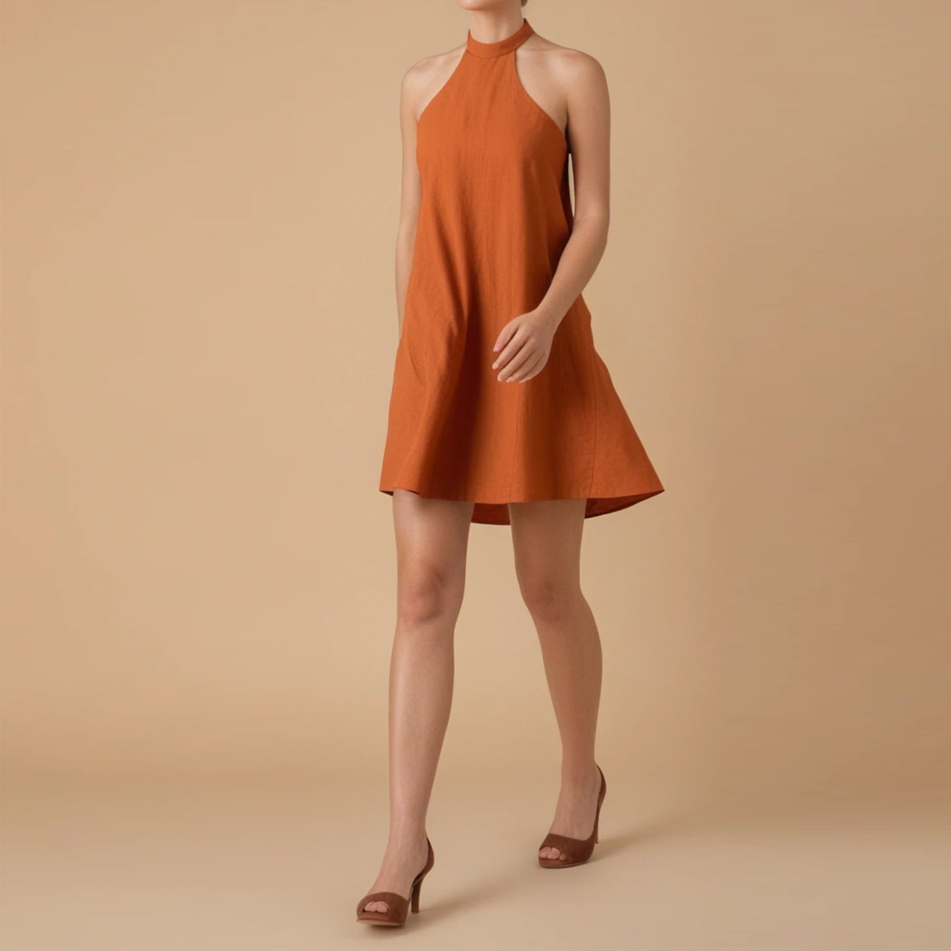 A woman models a sleeveless, burnt orange halter-neck summer dress made from lightweight fabric. The dress has a high neckline, flared A-line silhouette, and a slightly longer back hem. She pairs the dress with brown high-heeled sandals. The background is a solid beige color, giving the image a minimal and elegant look. This dress design is featured as part of a sewing pattern PDF.